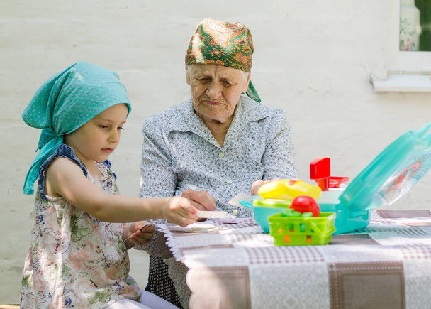 Ein kleines Mädchen spielt mit einer älteren Frau in einem Dorf auf dem Hof ein Brettspiel. Gemeinsam Spaß haben. Familienbeziehung zu Großmutter und Enkelin. - Foto, Bild