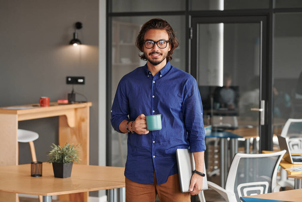 Portrait of smiling young mixed race app developer holding laptop and drinking coffee in coworking space - Φωτογραφία, εικόνα