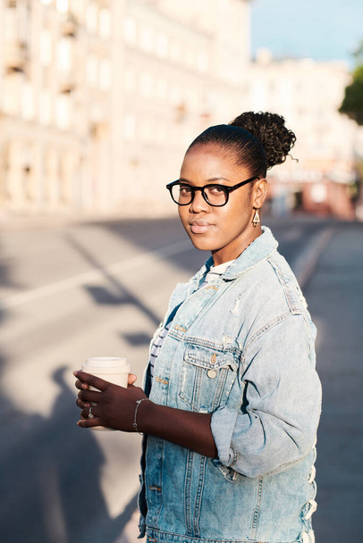 Portrait of content young Black woman in eyeglasses wearing denim jacket holding takeout coffee against cityscape - Photo, Image