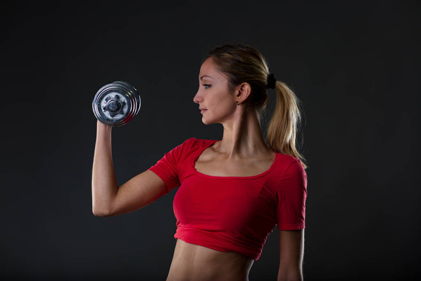 Rubia apasionada del fitness, levantando peso, enfatizando su estómago en forma. Camiseta de gimnasio roja, micro pantalones negros. Fondo negro borroso - Foto, Imagen