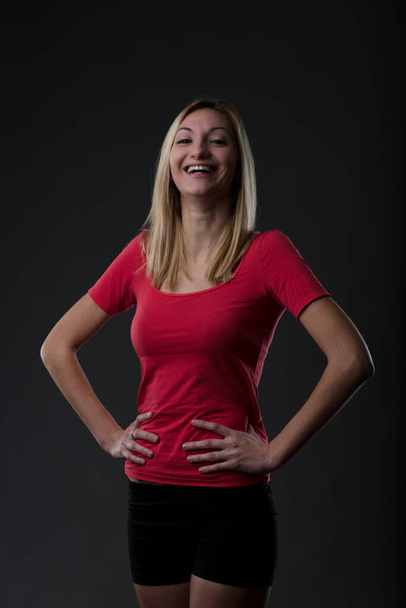 Blonde woman laughing with hands on hips. Portrait in gym attire, red shirt, and shorts. She laughs at those who tell her to accept herself as she is because she knows she wouldn't want to be with som - Photo, Image