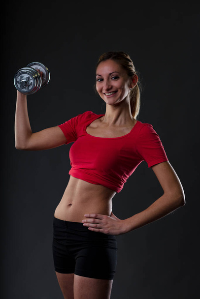 Fitness-leidenschaftliche Blondine, hebt Gewicht und betont ihren fitten Bauch. Rotes Turntop, schwarze Mikrohose. Unscharfer schwarzer Hintergrund - Foto, Bild