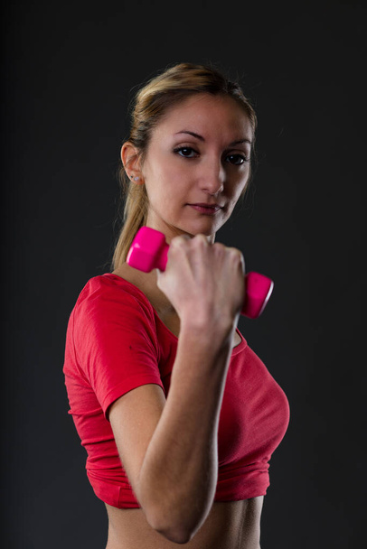 Vrouw tilt roze halter, wazige achtergrond. Blond, rood topje, vlakke buik, trots op fitness doelen. Volgend Mens Sana In Corpore Sano motto - Foto, afbeelding