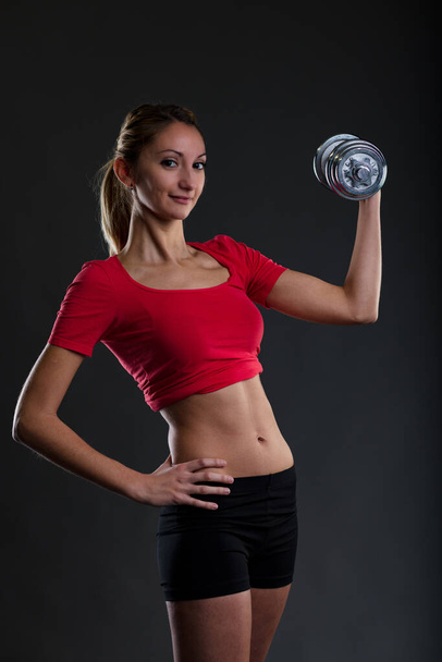 Blonde girl lifting weights in the gym, showcasing her flat stomach. Wearing a red gym top, black micro pants. Passionate about fitness! - Foto, afbeelding