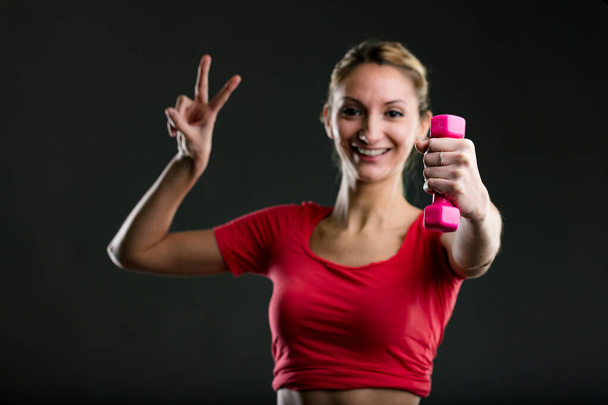 Woman lifting pink dumbbell, giving V-sign, smiling. Blonde, red top, flaunts flat stomach, proud of fitness goals. Her motto: 'Smart in a sexy body, complain less' - Photo, Image