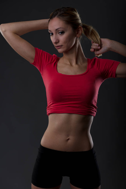 Fit woman maintains toned body with gym workouts. Physical activity reflects determination and responsibility. Portrait of blonde girl in red shirt, flat belly, black shorts against black background - Foto, imagen