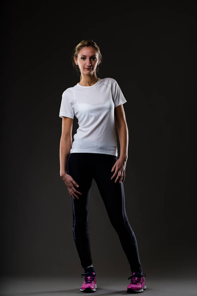 Blonde woman ready for workout, standing comfortably, hands on hips, looking at the camera. On black background, wearing dark tight pants, fuchsia sneakers, and a comfortable white t-shirt - Foto, imagen