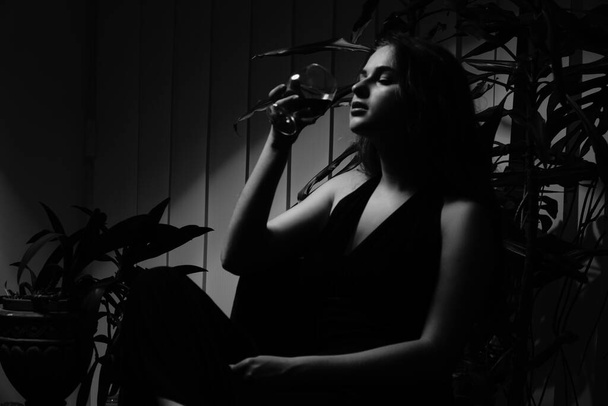 ασπρόμαυρη φωτογραφία μιας όμορφης αισθησιακής γυναίκας με μαύρο φόρεμα με ένα ποτήρι ουίσκι που ποζάρει για την κάμερα - Φωτογραφία, εικόνα