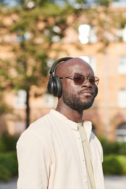 Portret van een serieuze zwarte man in een zonnebril en een draadloze koptelefoon die alleen in de stad loopt - Foto, afbeelding