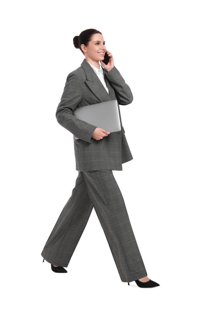 Beautiful happy businesswoman with laptop talking on smartphone while walking against white background - Photo, Image