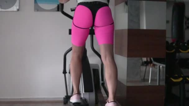 Una donna si allena su un simulatore di ellisse in una palestra coperta, un primo piano di gambe gonfie in pantaloncini rosa sexy sottolineando la figura. - Filmati, video