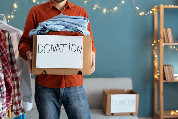 Caring male volunteer holding donation box full of clothes to needy people. Man preparing things in package donate to poor, those in need. Reuse, recycle, charity concept. copy space - 写真・画像