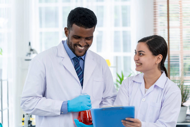 Young male scientist and girl friend doing experiment with liquid in beaker and checking in paperwork on clipboard in science lab, They are discussion with happy, copy space - Photo, image