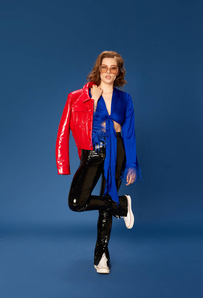 Portrait of beautiful young woman in stylish clothes, top, jacket and leather pants standing against blue studio background. Concept of human emotions, female fashion, beauty, party, lifestyle - Foto, Imagem