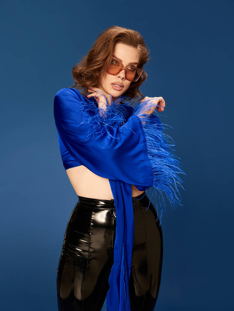 Portrait of attractive young brunette woman in trendy sunglasses, stylish top and leather pants posing over blue studio background. Concept of human emotions, female fashion, beauty, party, lifestyle - Photo, Image