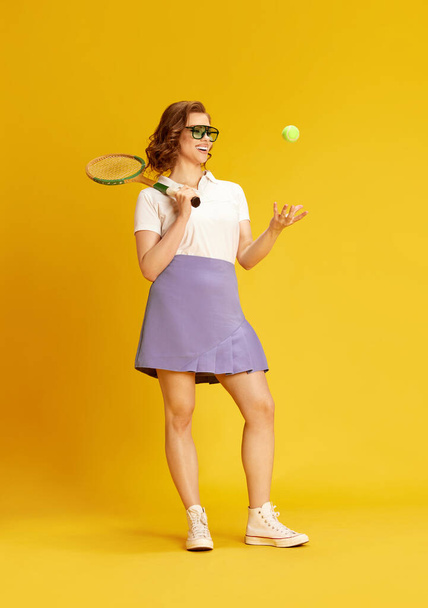 Portrait of young, beautiful girl in sportswear and glasses, smiling posing with tennis racket and ball against yellow studio background. Concept of sport, active lifestyle, emotions, fashion, hobby - Foto, afbeelding