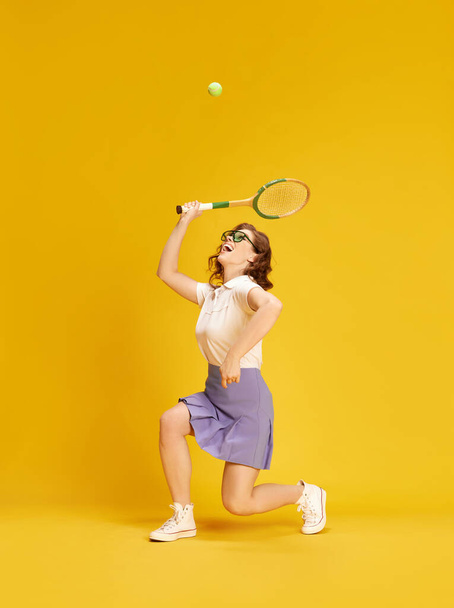 Portrait of young, active, pretty girl in sportswear, tennis playing in motion hitting ball with racket against yellow studio background. Concept of sport, active lifestyle, emotions, fashion, hobby - Φωτογραφία, εικόνα