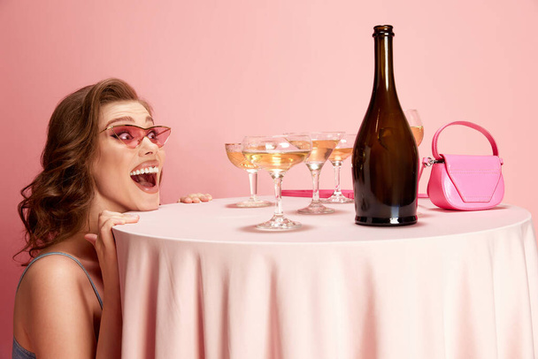 Portrait of young beautiful woman peeking out table served with champagne against pink studio background. Excitement and happiness. Concept of emotions, celebration, party, fashion - Foto, Imagen
