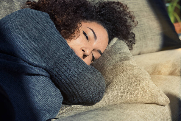 Sunlight graces a beautiful girl with closed eyes on her couch. Curly-haired, wrapped in her warm sweater and home's comfort, she may be dreaming, resting or asleep. She's simply beautiful - Photo, Image