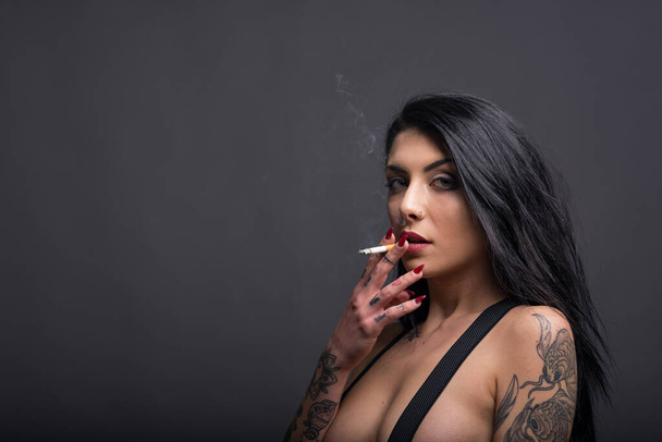 Sensual portrait of beautiful woman with tattoo smoking against dark background. - Photo, Image