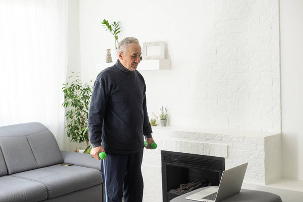 Sport in mature age. Happy middle-aged man doing stretching exercises in front of laptop at home, watching online tutorials, in living room interior, free space. - Foto, Imagen