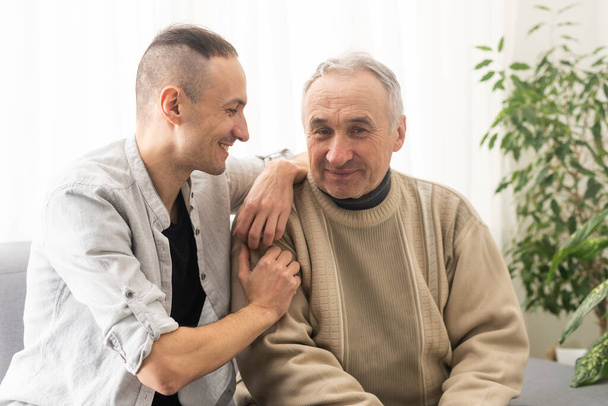 Young adult caucasian son listening and supporting his old elderly senior father at home indoors. Care and love concept - Foto, Bild