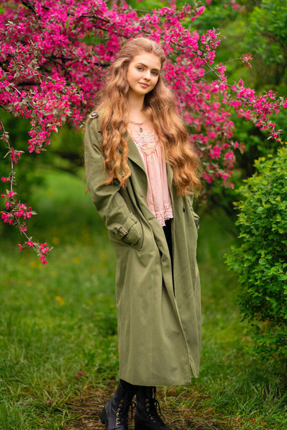 Chica rubia bastante joven con el pelo largo rizado en blusa de encaje vintage y capa verde de pie en el parque de primavera cerca de flores de flor rosa. Ternura modelo romántico posando y mirando a la cámara. - Foto, Imagen