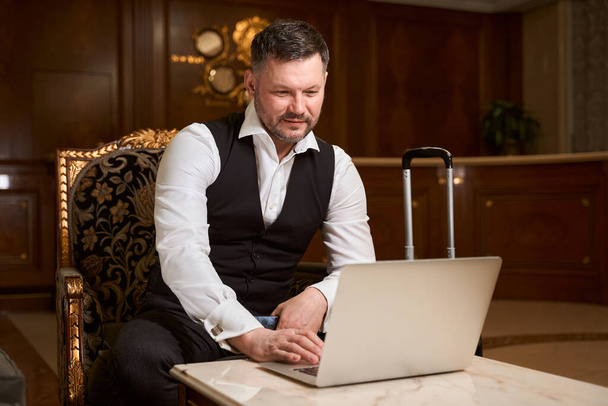 Handsome elegant businessman in formal suit sitting on armchair and using laptop at table near suitcase in hotel lobby - Foto, imagen