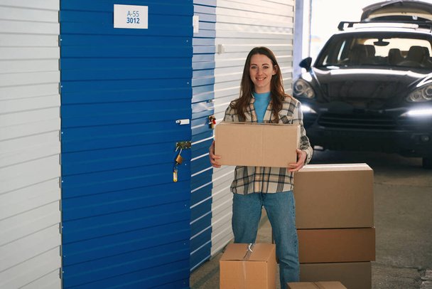 Smiling lady is holding a large box near open door of the pantry in the storage warehouse. In the background is standing a car with open trunk - Fotó, kép