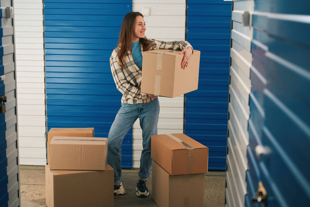 Young lady is holding a big cardboard box and standing in storage warehouse with many boxes around - Foto, Imagem