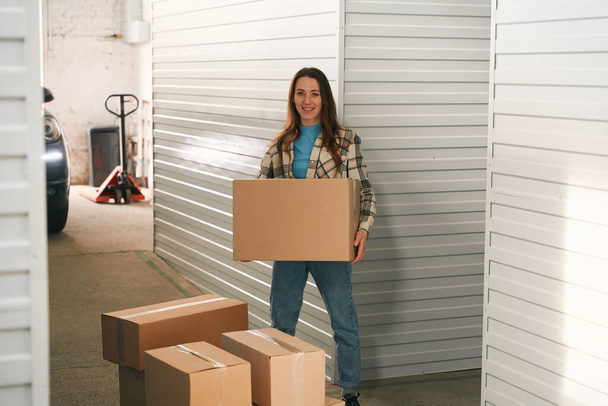 Happy woman is holding a big cardboard box and standing in storage warehouse with many boxes around - Foto, Bild
