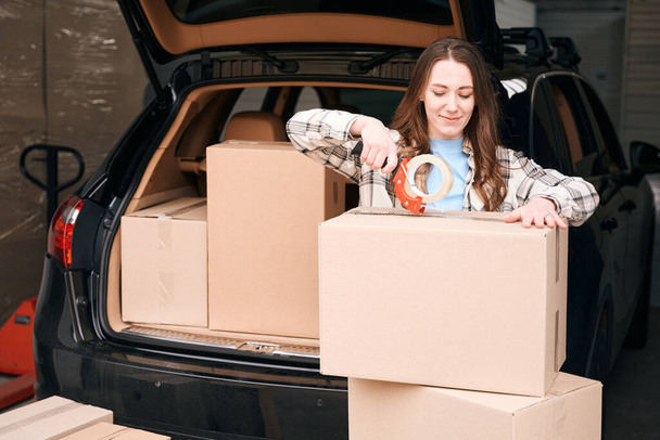 Cute lady is packing cardboard boxes into the trunk of car in a storage warehouse - 写真・画像