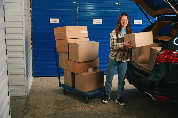 Smiling woman is puting cardboard boxes in the trunk of a car into warehouse with self storage unit - Zdjęcie, obraz