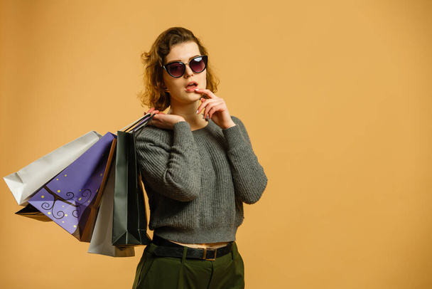 Portrait of an excited beautiful girl wearing sunglasses holding shopping bags isolated over beige background. - Zdjęcie, obraz