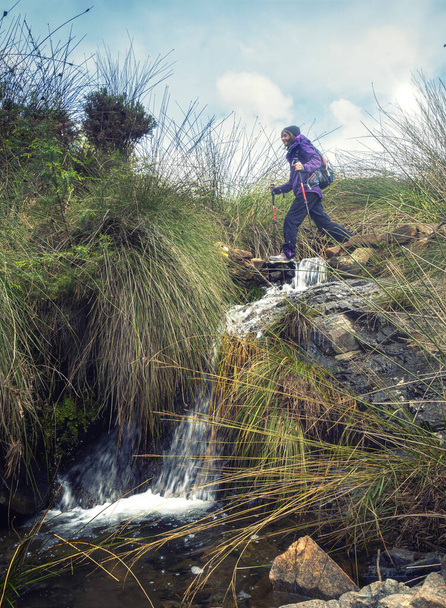 Женщина одна с тростью и рюкзаком, пересекающими ручей. Прогулка среди природы с водопадом. Свежесть и приключения. Копирование пространства. - Фото, изображение