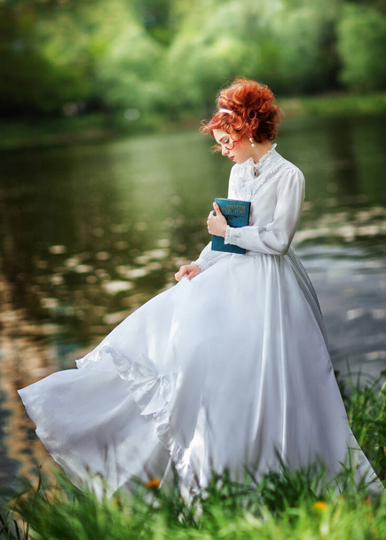Beautiful red haired girl in white long vintage dress standing near river with book.Art work of romantic woman with  lace umbrella.  - Фото, зображення