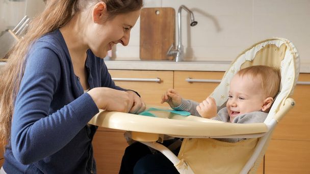 Happy laughing mother with baby son sitting in highchair on kitchen. - 写真・画像
