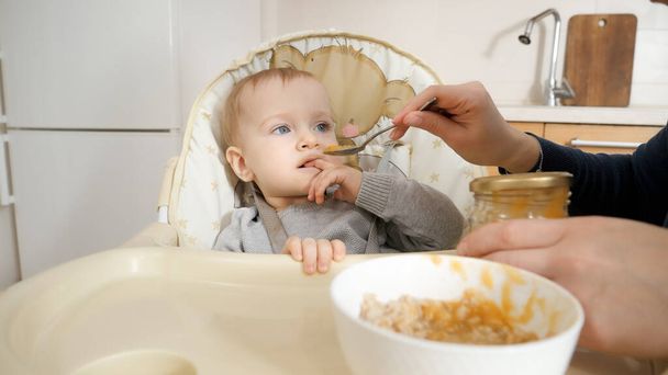 Schattige kleine jongen glimlacht naar zijn moeder terwijl ze hem voedt met pap. Begrip ouderschap, gezonde voeding en babyvoeding - Foto, afbeelding