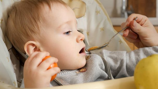 Closeup portrait of little baby boy getting messy while mother is feeding him from spoon. Concept of parenting, healthy nutrition and baby feeding - Photo, Image