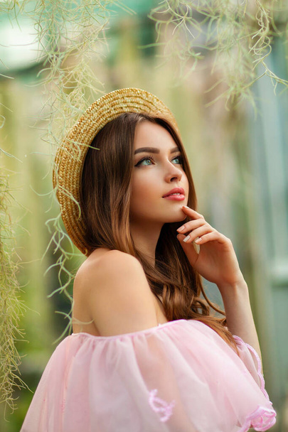 Schöne Mädchen in rosa Vintage-Kleid und Strohhut steht auf grünem Hintergrund mit Pflanzen. Kunstwerk der romantischen Frau. Hübsche Zärtlichkeit Modell posiert und schaut in die Kamera. - Foto, Bild