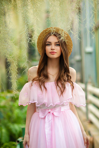 Krásná dívka v růžové retro šaty a slaměný klobouk stojící na zeleném pozadí s rostlinami. Umělecké dílo romantické ženy.Pretty něha model pózování a pohled do kamery. - Fotografie, Obrázek