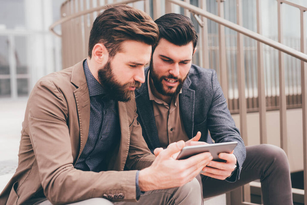 Два елегантних сучасного бородатого бізнесмена на відкритому повітрі, що сидить на сходах, обмінюючись планшетом, віддалено обговорюють мозковий штурм
 - Фото, зображення