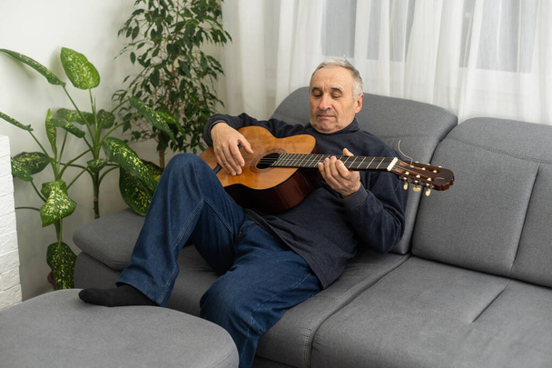 先輩がギターを弾いています。ソファーに座ってギターを弾いている老人。セーターを再生することを学ぶの白髪の成熟した男の肖像画。引退生活を自宅で楽しむ - 写真・画像