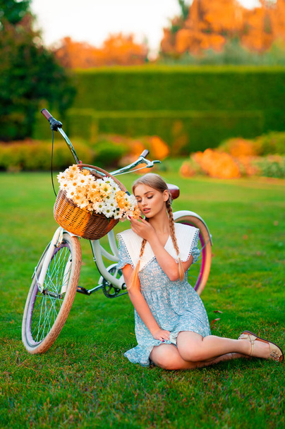 Beautiful young blonde smiling girl with short blue dress sitting near white bicycle with basket of huge bouquet of chamomiles.Summer lady enjoying the nature outdoor.Green background with flowers. - Zdjęcie, obraz