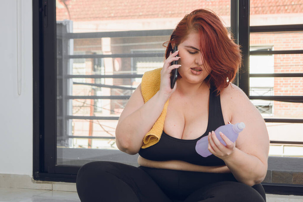 young plus size woman in sportswear sitting talking on the phone reading the label of a bottle of water, before doing stretching exercises - Photo, Image