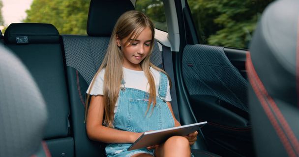 Attaractive little girl passenger in car reading digital tablet and watching cartoons online. Child networking with tablet computer while travelling by car on summer vacation. - Фото, зображення