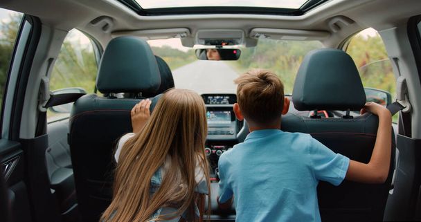 Back view. Kids looking outside and pretty woman riding by road wheel. Child boy and girl cheerful parent spending time together in car. - Photo, image