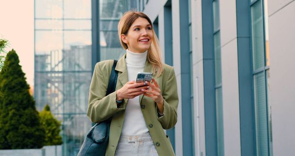 Fröhliche junge Kaukasierin, die mit dem Handy in der Hand auf der Straße zum Büro läuft. Lächelnde Dame mit Handy in der Hand draußen - Foto, Bild