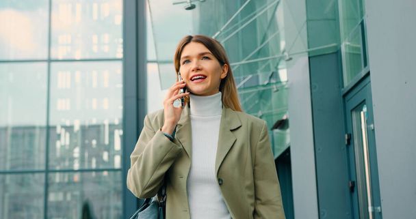 Caucasian woman speaking on mobile phone walking beside office outdoors - Photo, Image
