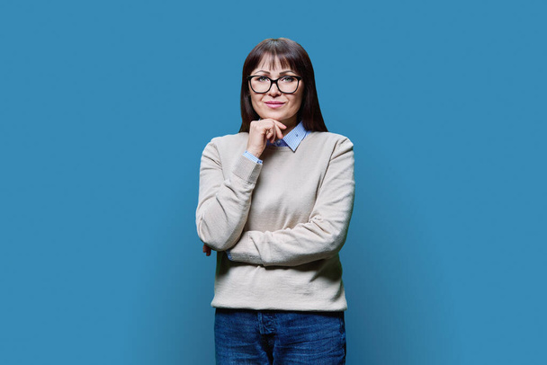 Portrait of smiling middle aged brunette woman wearing glasses in jumper looking at camera, on blue background. Mature middle age, professionalism, service, health beauty care expert - Фото, изображение